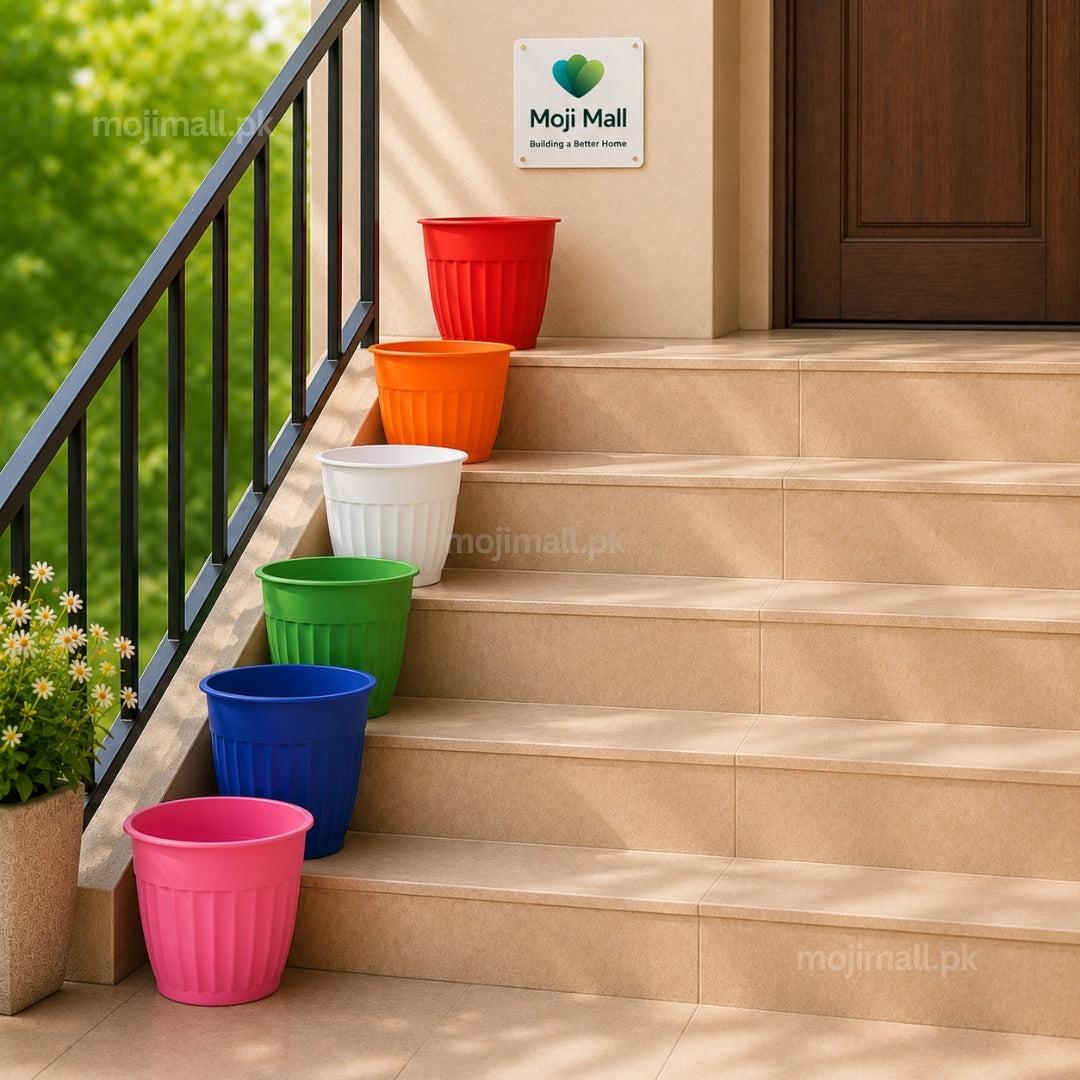 Six colorful plastic flower pots in pink, blue, green, white, orange, and red are arranged on outdoor concrete stairs next to a black metal railing, with a Moji Mall sign on the wall above.
