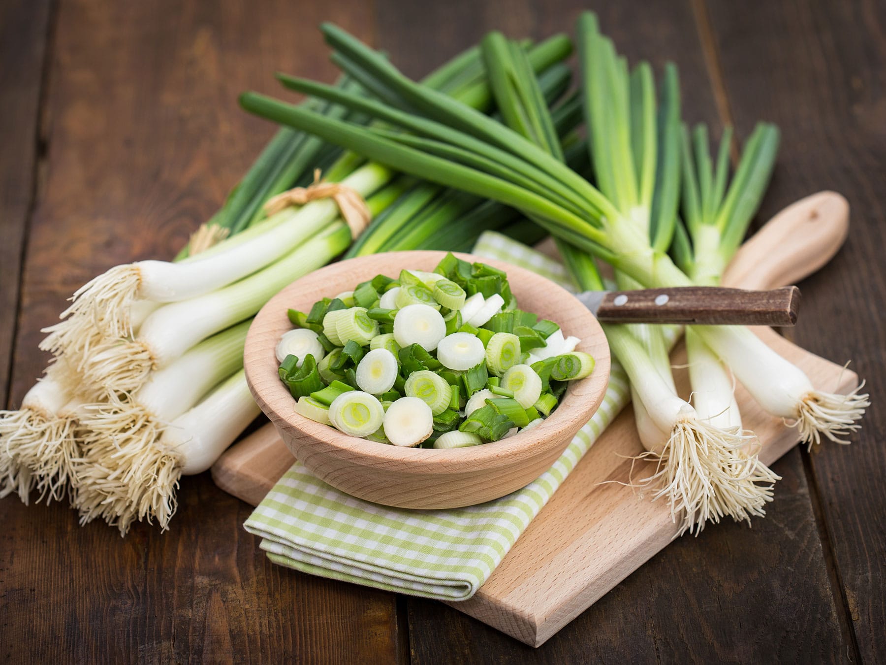 Green onions with a bowl of chopped green onions on a wooden cutting board.